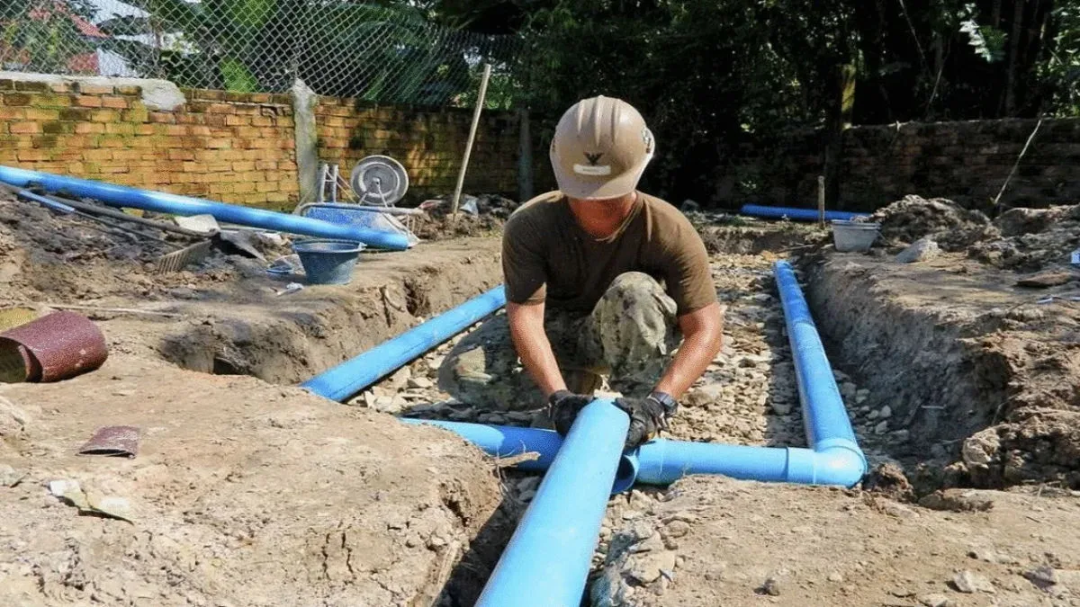 A worker in a hard hat carefully aligns blue PVC piping within a freshly excavated ground trench.