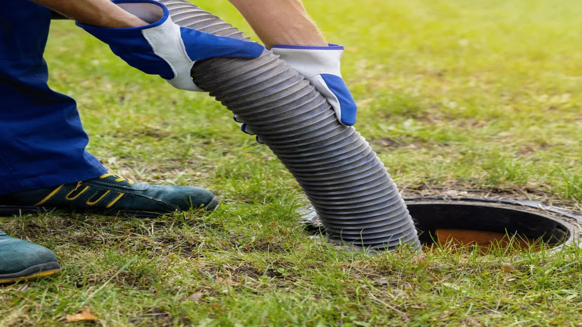 A worker wearing protective blue gloves carefully guides a flexible vacuum hose into a residential drain.