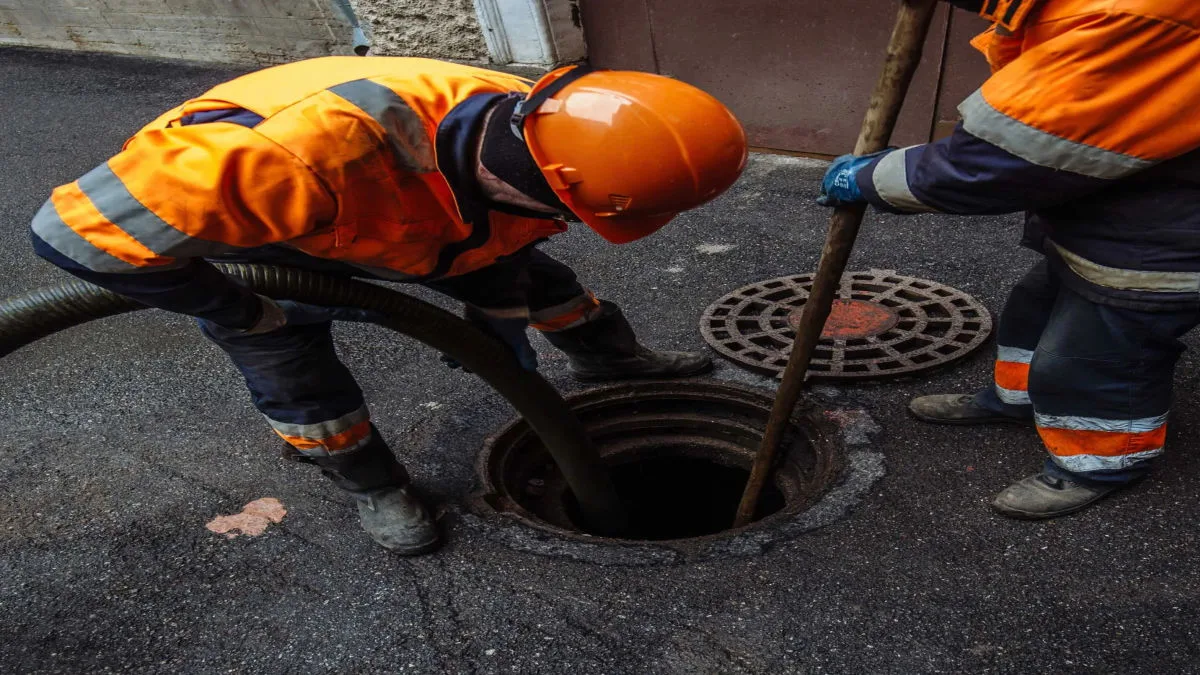 A technician wearing orange safety gloves guides a heavy-duty suction pipe into an open drain.